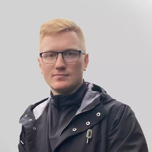 Portretinė nuotrauka – jaunas vyras trumpais šviesiais plaukais, vilkintis juodą striukę su pilku pašiltinimu. Jis žiūri į kamerą su rimta veido išraiška, fonas neutralus, pilkšvas.